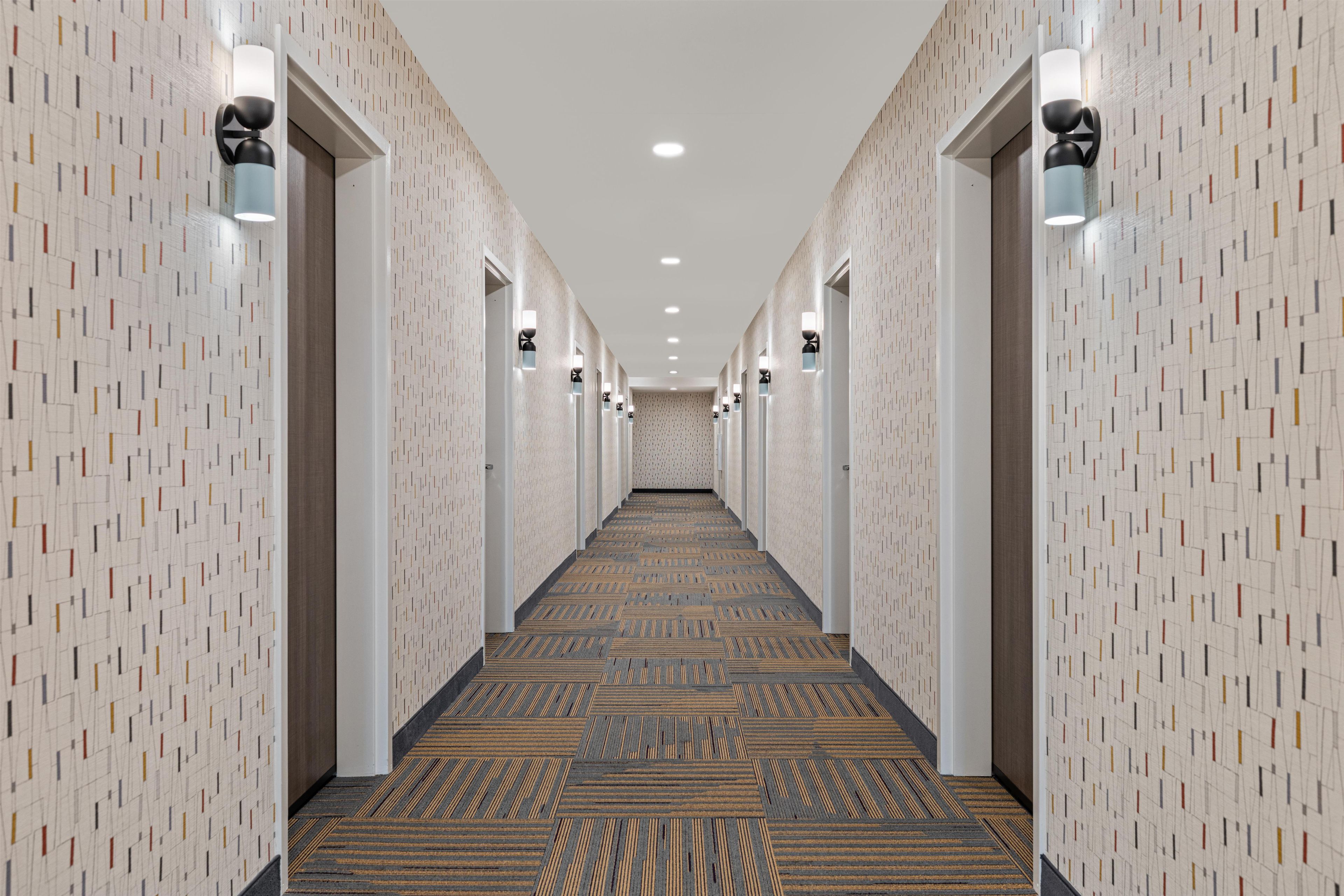 A hotel hallway with patterned walls, recessed lighting, and a carpeted floor.
