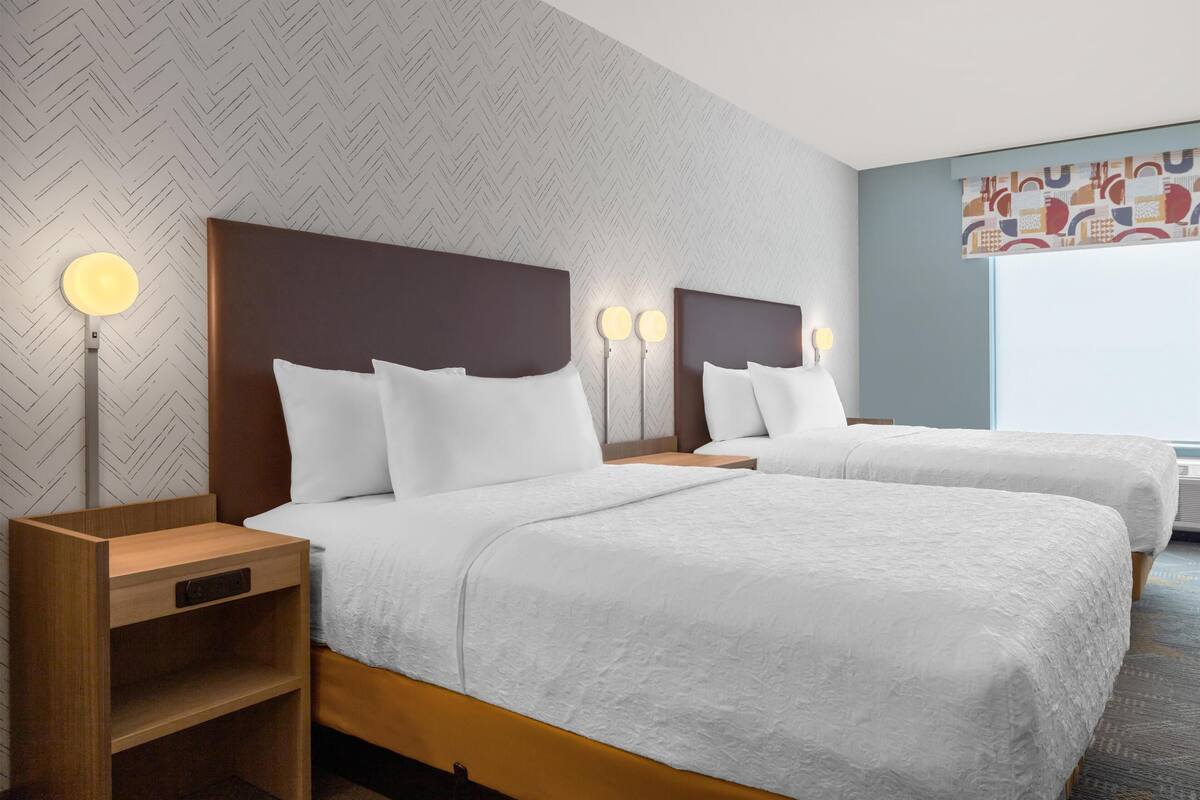 A hotel room with two beds, a wooden nightstand, a wall-mounted lamp, and a patterned wall.
