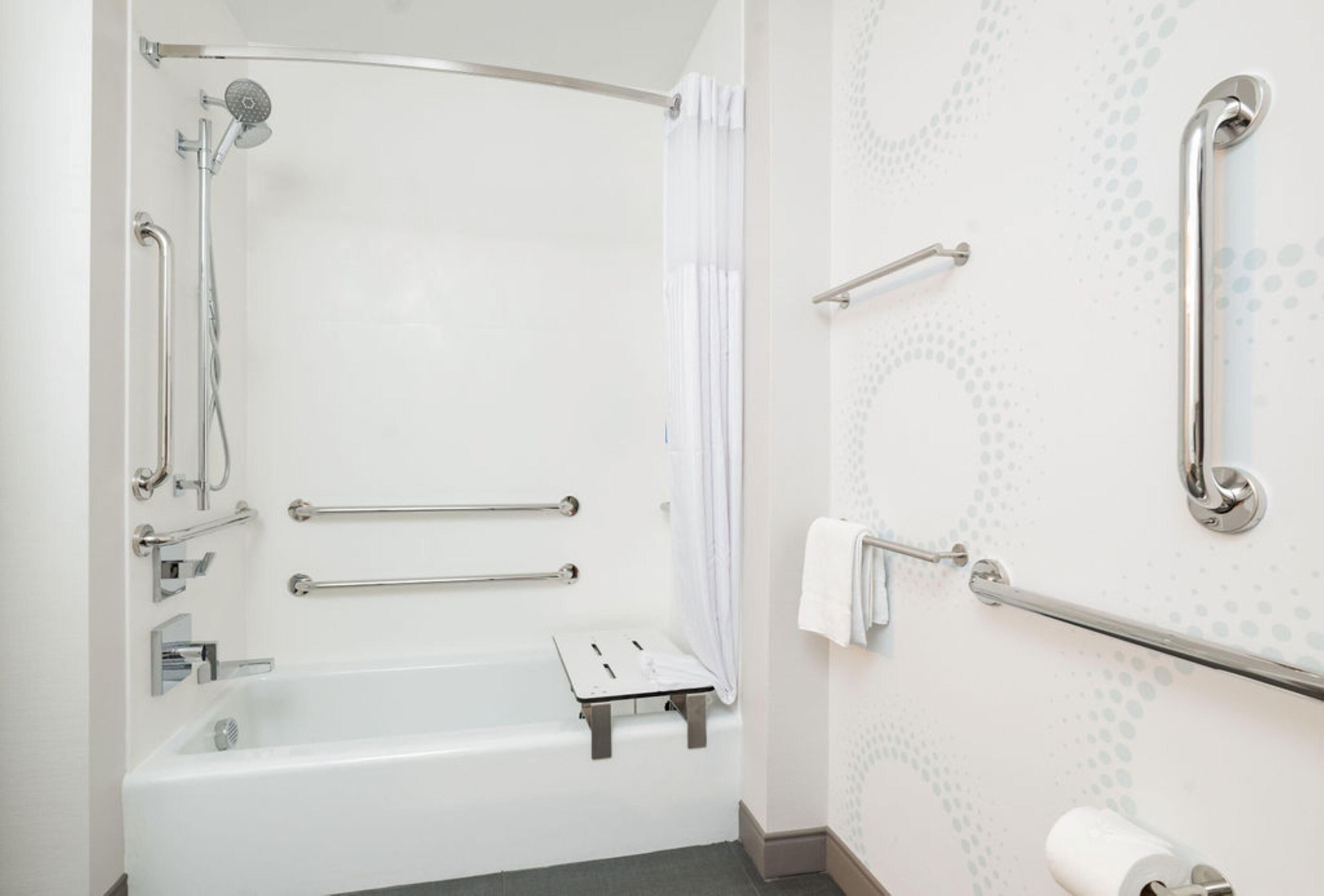 Un baño con una bañera blanca, barras de apoyo, cabezal de ducha y un dispensador de papel higiénico.