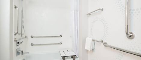 Un baño con una bañera blanca, barras de apoyo, cabezal de ducha y un dispensador de papel higiénico.