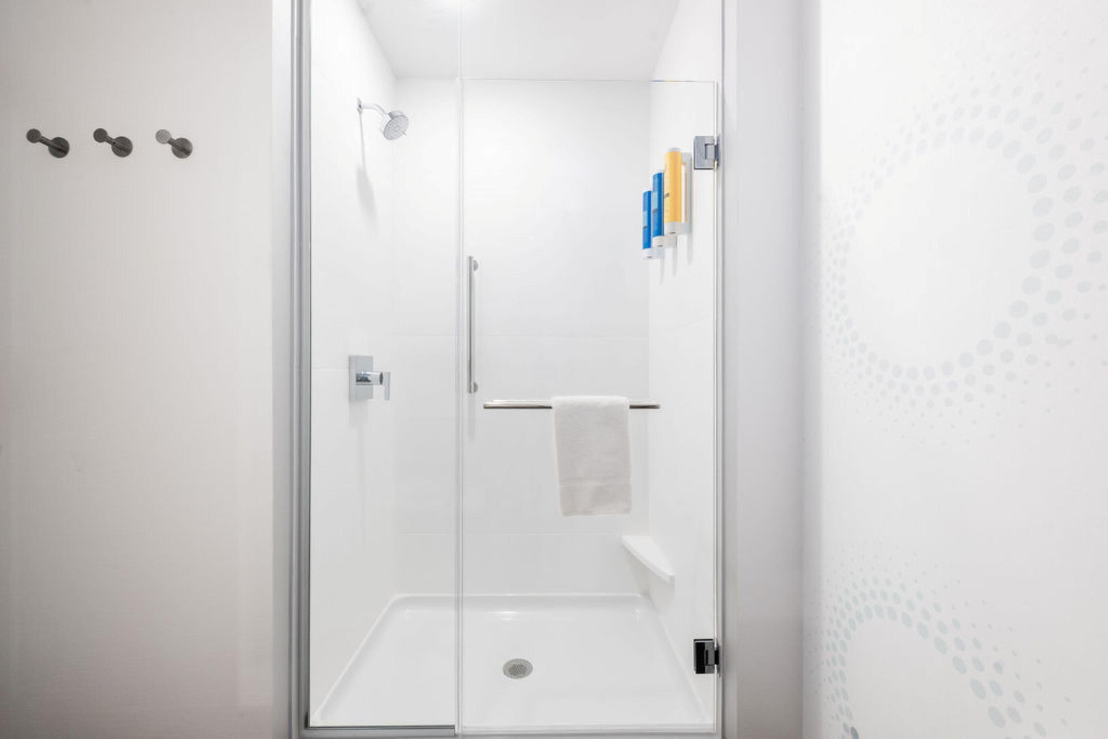 A glass-enclosed shower stall with a white towel hanging on a rack, a showerhead, and a small shelf with toiletries.