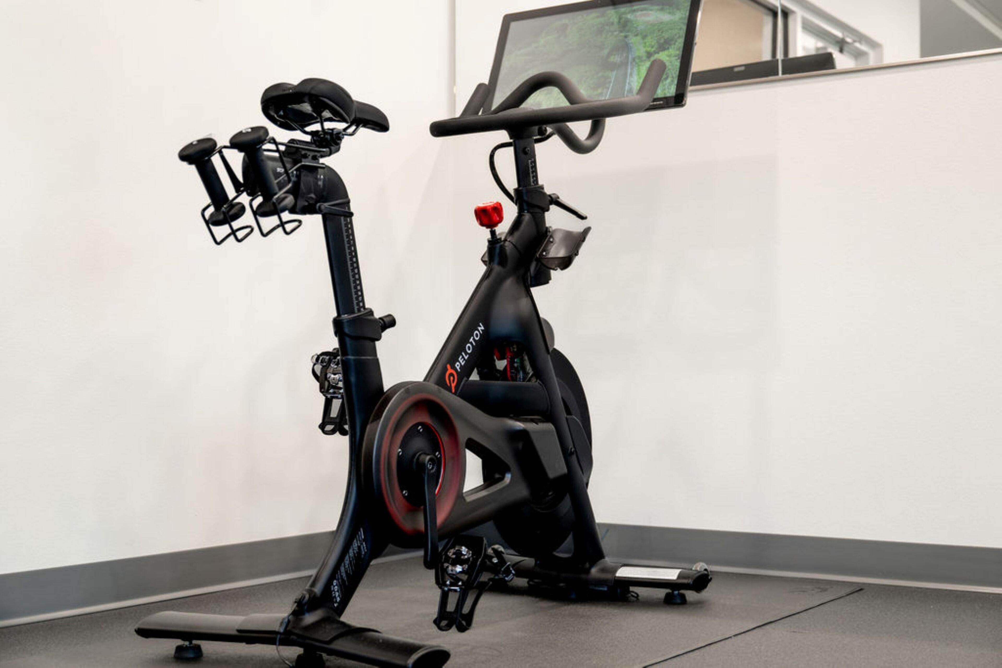 A stationary exercise bike with a screen and handlebars, positioned in a room with a plain wall and a window.