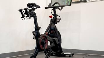 A stationary exercise bike with a screen and handlebars, positioned in a room with a plain wall and a window.