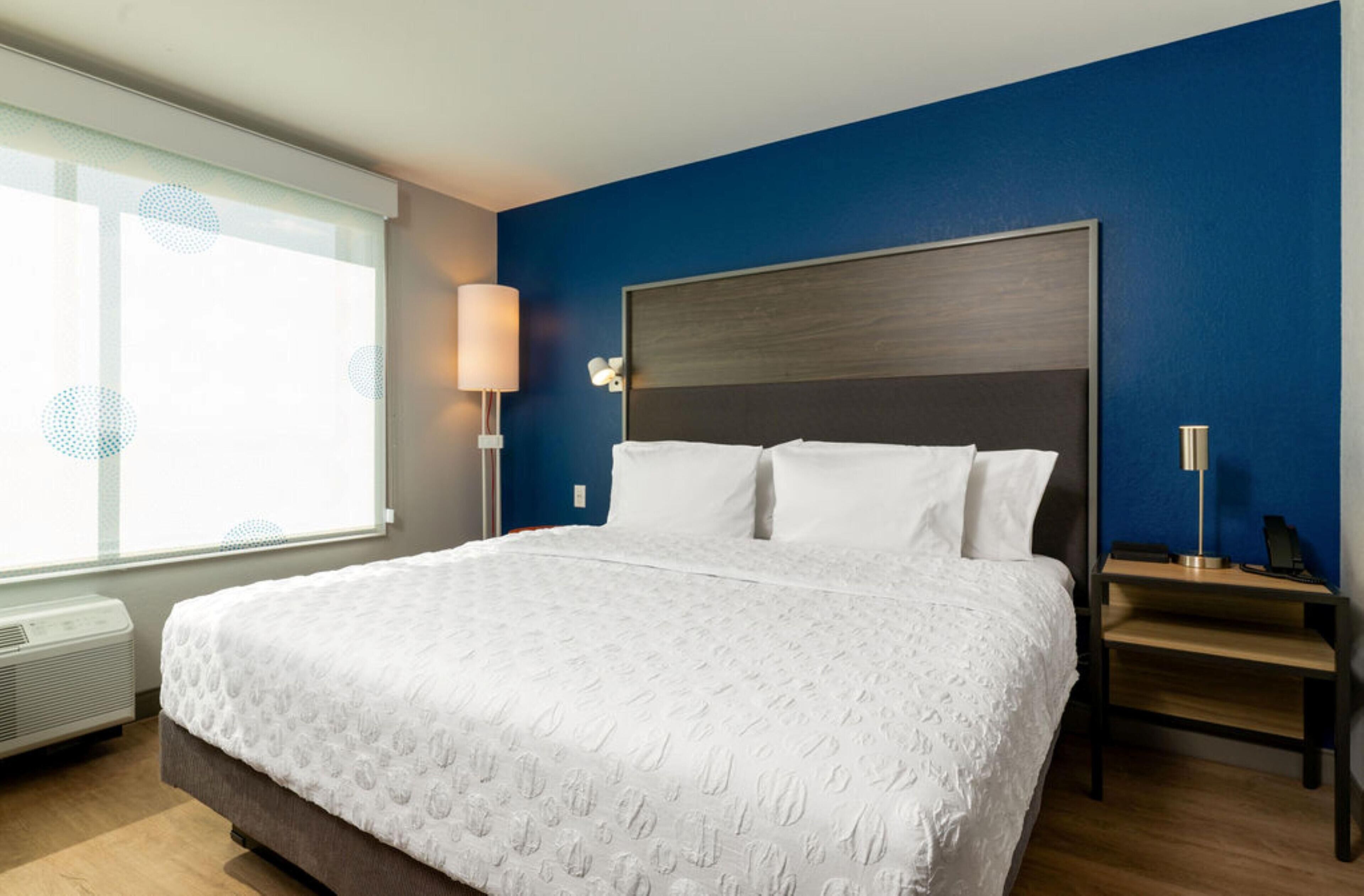 A hotel room with a large bed, a bedside table, a lamp, and a window with blinds.