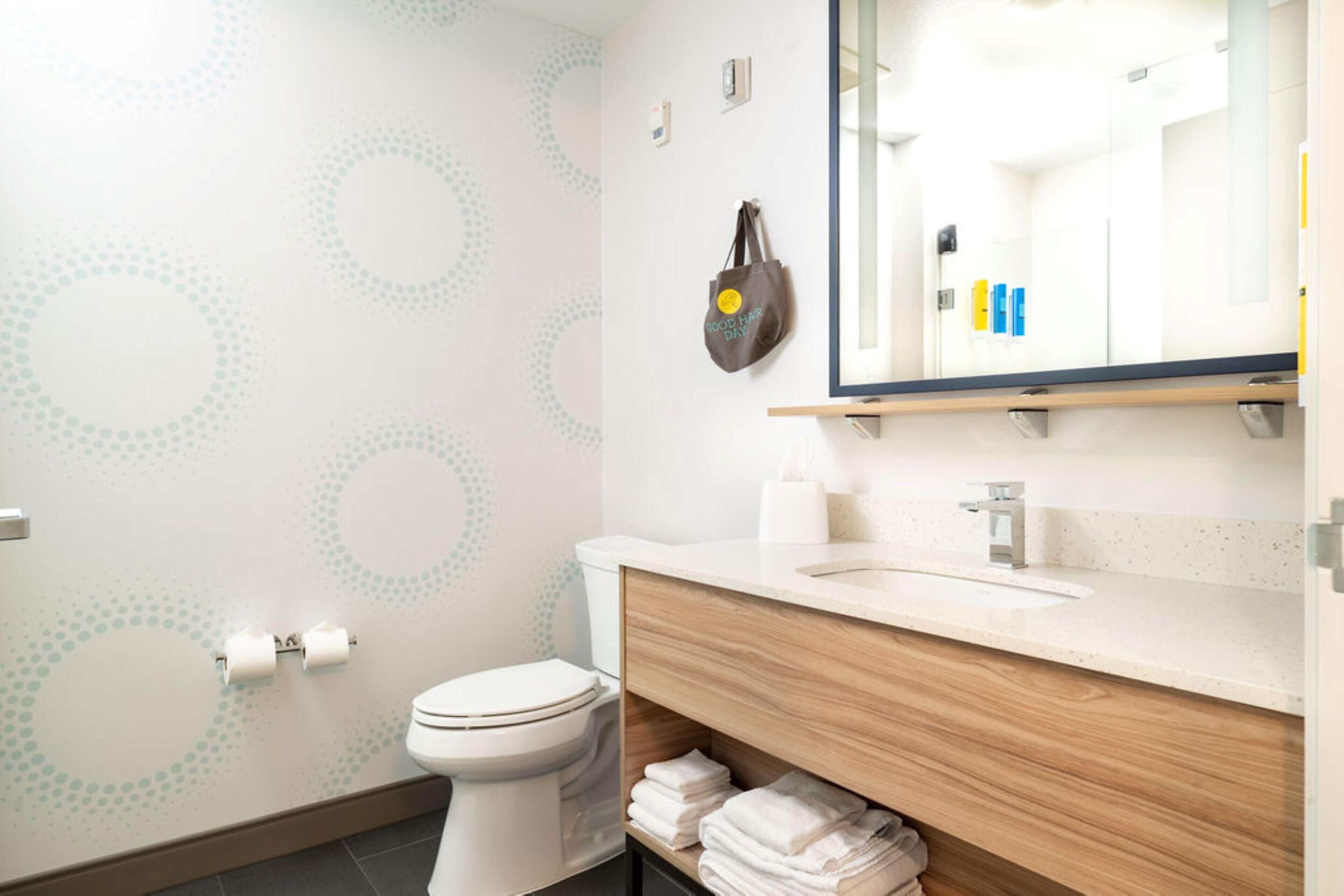 Un baño moderno con inodoro, lavamanos y espejo. Hay toallas apiladas en un estante y un bolso colgado en la pared.