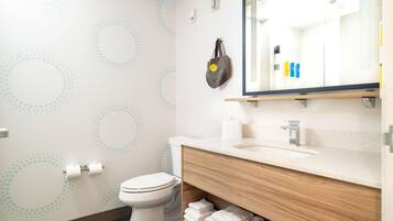 Un baño moderno con inodoro, lavamanos y espejo. Hay toallas apiladas en un estante y un bolso colgado en la pared.
