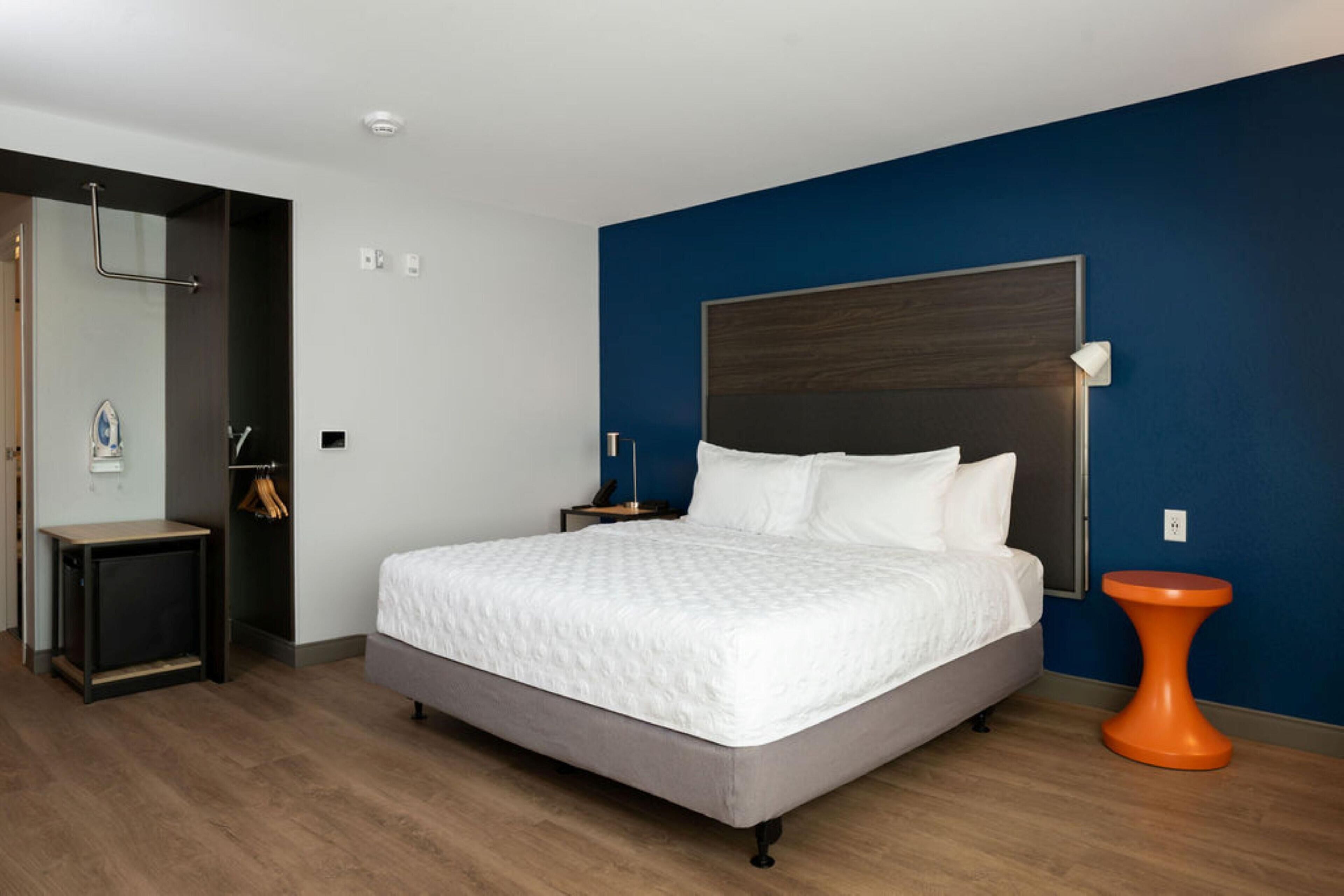 Une chambre d’hôtel moderne avec un grand lit, un plancher en bois, un mur d’accent bleu et une petite table d’appoint orange.