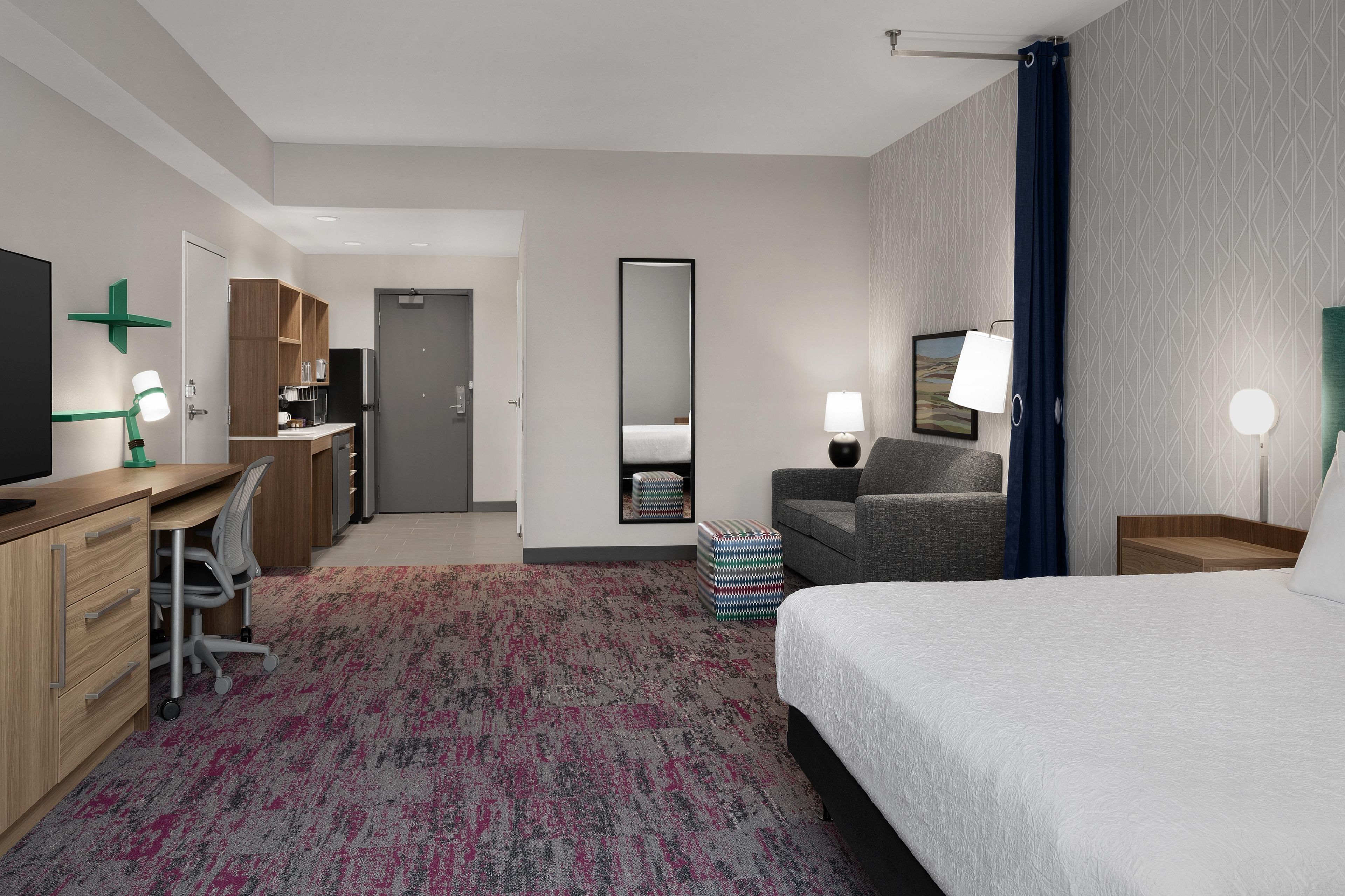 A hotel room with a bed, desk, chair, and a small kitchenette.