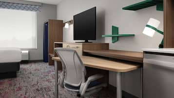 A hotel room with a bed, a desk with a computer, a chair, a TV, a desk lamp, and a window with curtains.