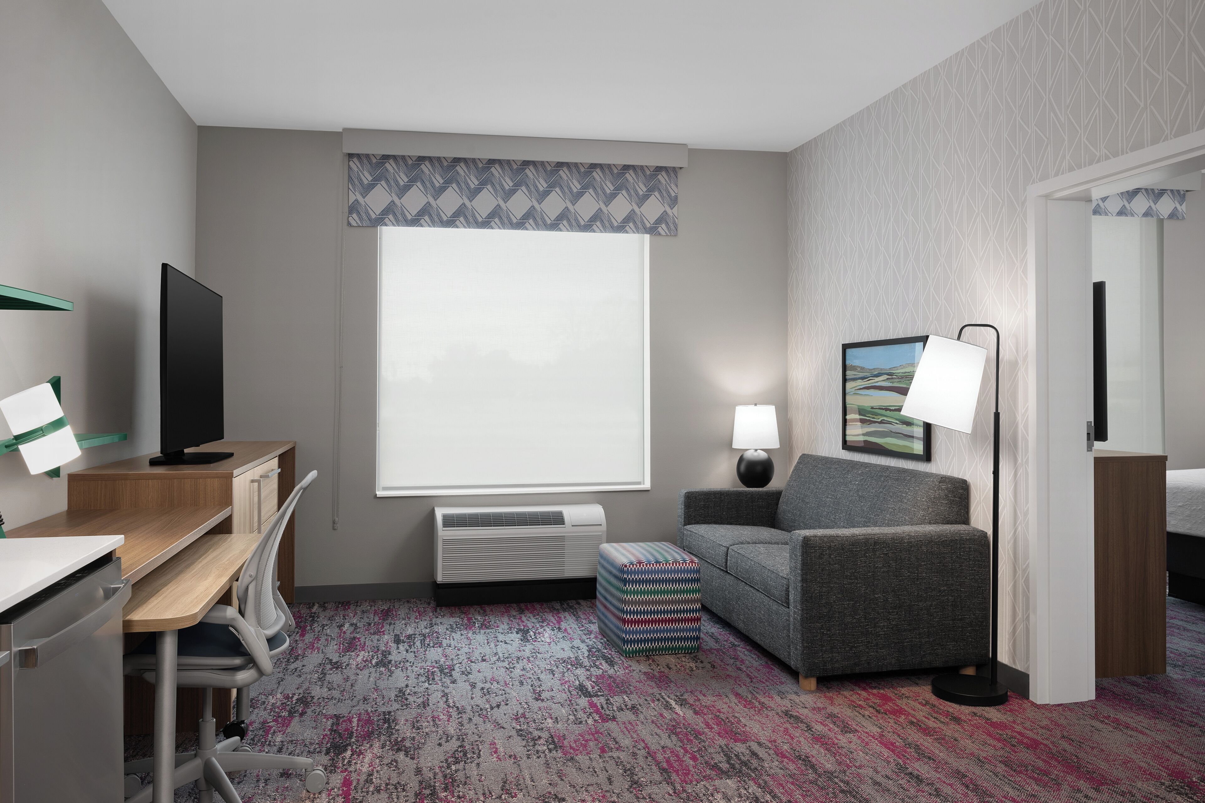 Habitación de hotel con escritorio, silla, televisor, sofá, lámpara y un ventanal grande.