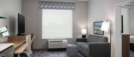 Habitación de hotel con escritorio, silla, televisor, sofá, lámpara y un ventanal grande.