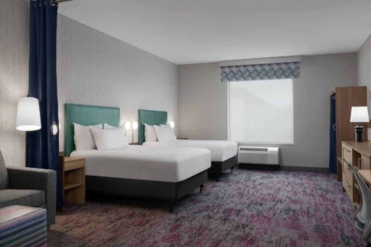 A hotel room with two beds, a desk, a chair, a window with a view, and a radiator.