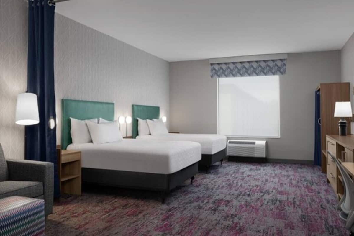 A hotel room with two beds, a desk, a chair, a window with a view, and a radiator.