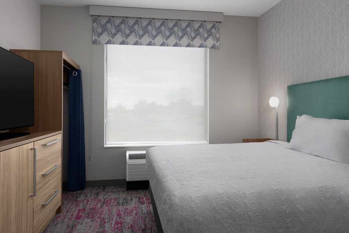A hotel room with a bed, a television on a wooden cabinet, a window with a patterned curtain, and a wall-mounted light.