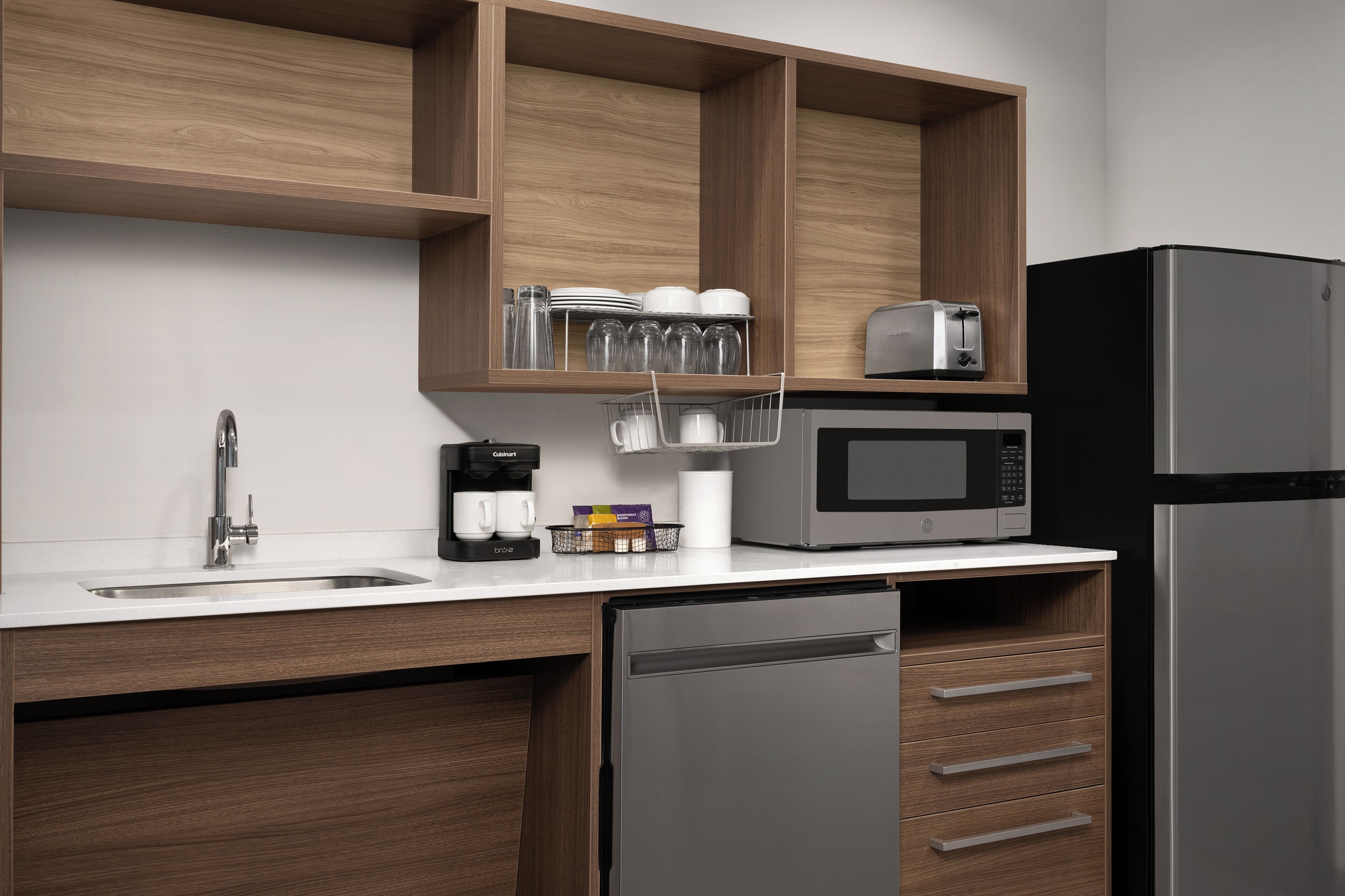 Une cuisine moderne dotée d’un réfrigérateur en acier inoxydable, d’un four à micro-ondes encastré, d’une machine à café et de meubles de rangement en bois.