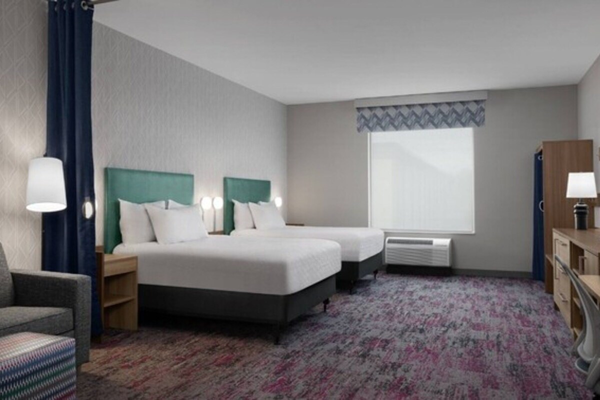 A hotel room with two beds, a desk, a chair, a window with a view, and a radiator.