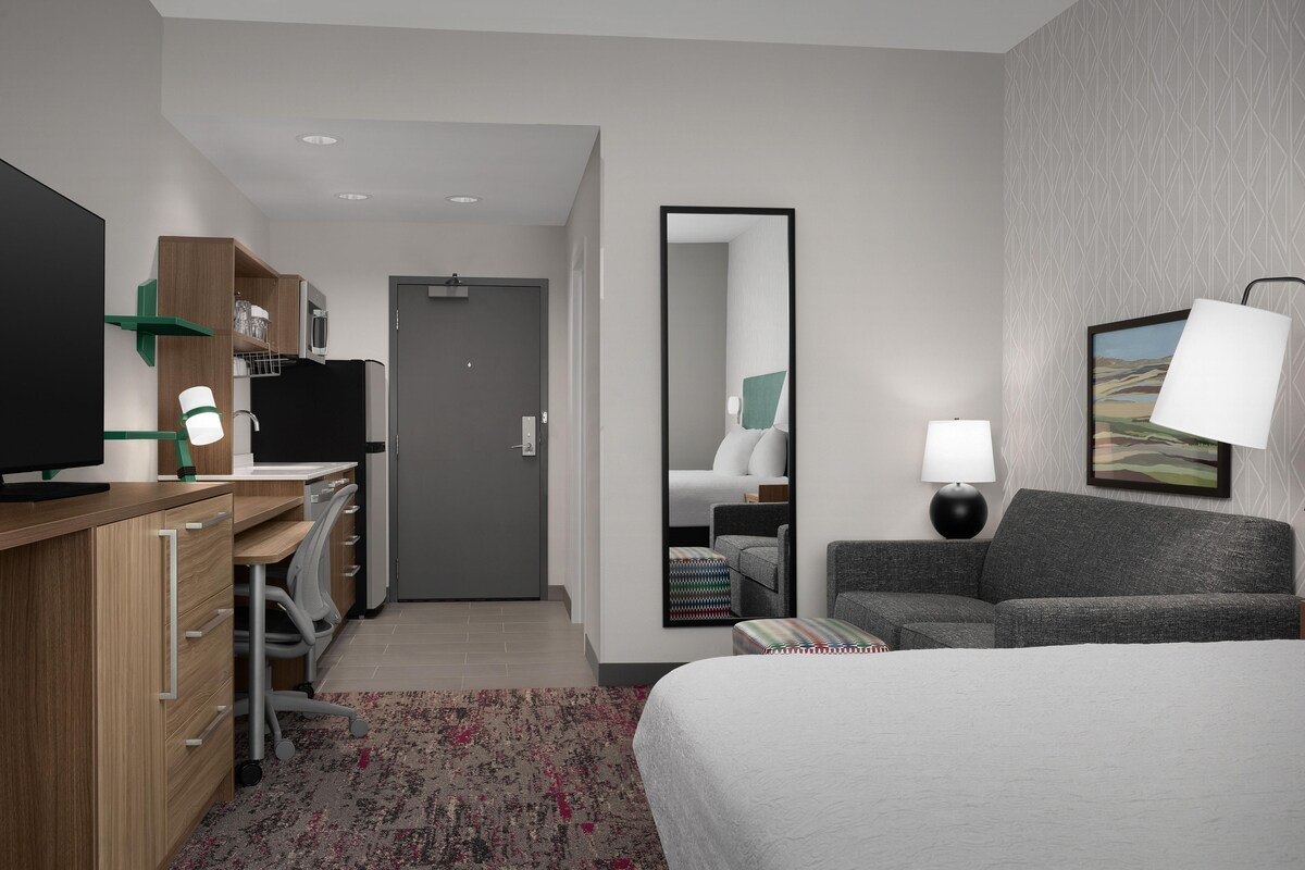 A hotel room with a bed, a desk with a computer, a chair, a sofa, a mirror, and a television.