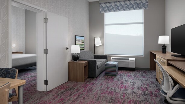 A hotel room with a bed, a desk, a chair, a small table, a lamp, a picture on the wall, and a window with a view.