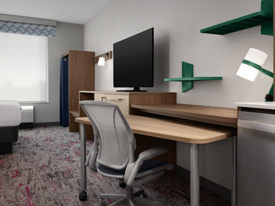 A hotel room with a bed, desk, chair, television, and a green wall-mounted shelf.