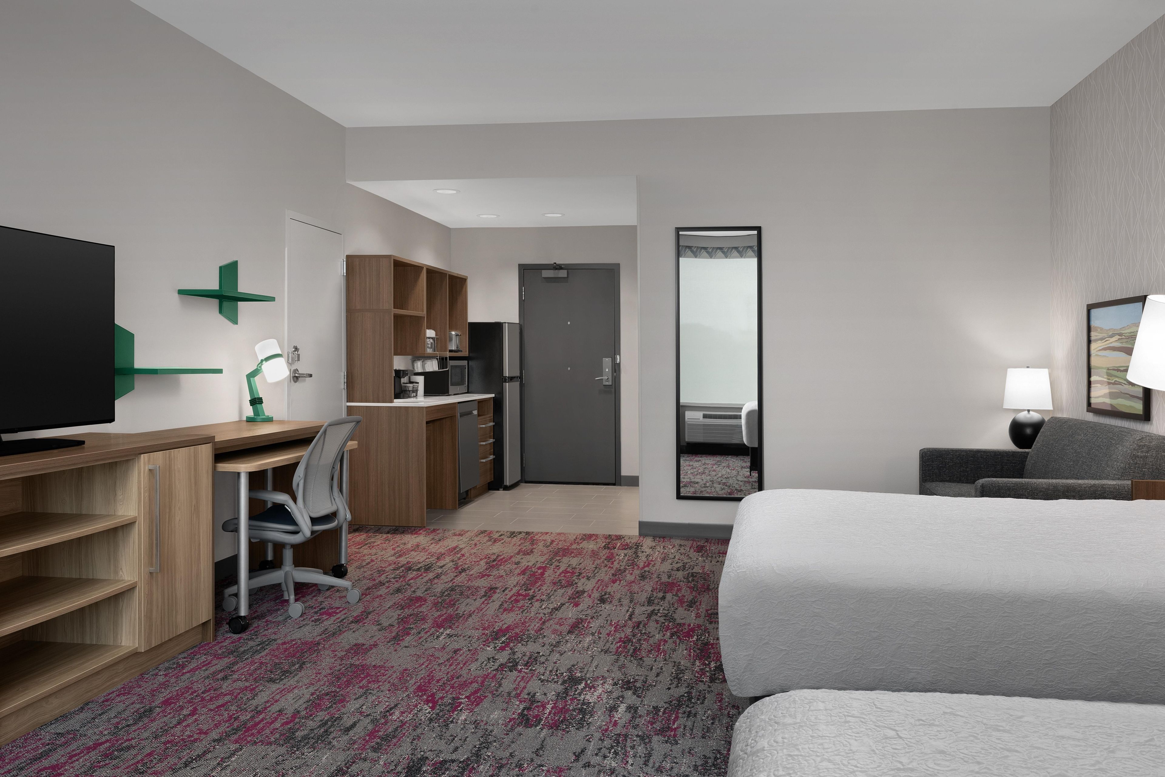 A hotel room with a bed, a desk with a chair, a television, a wardrobe, a kitchen area, and a bathroom.