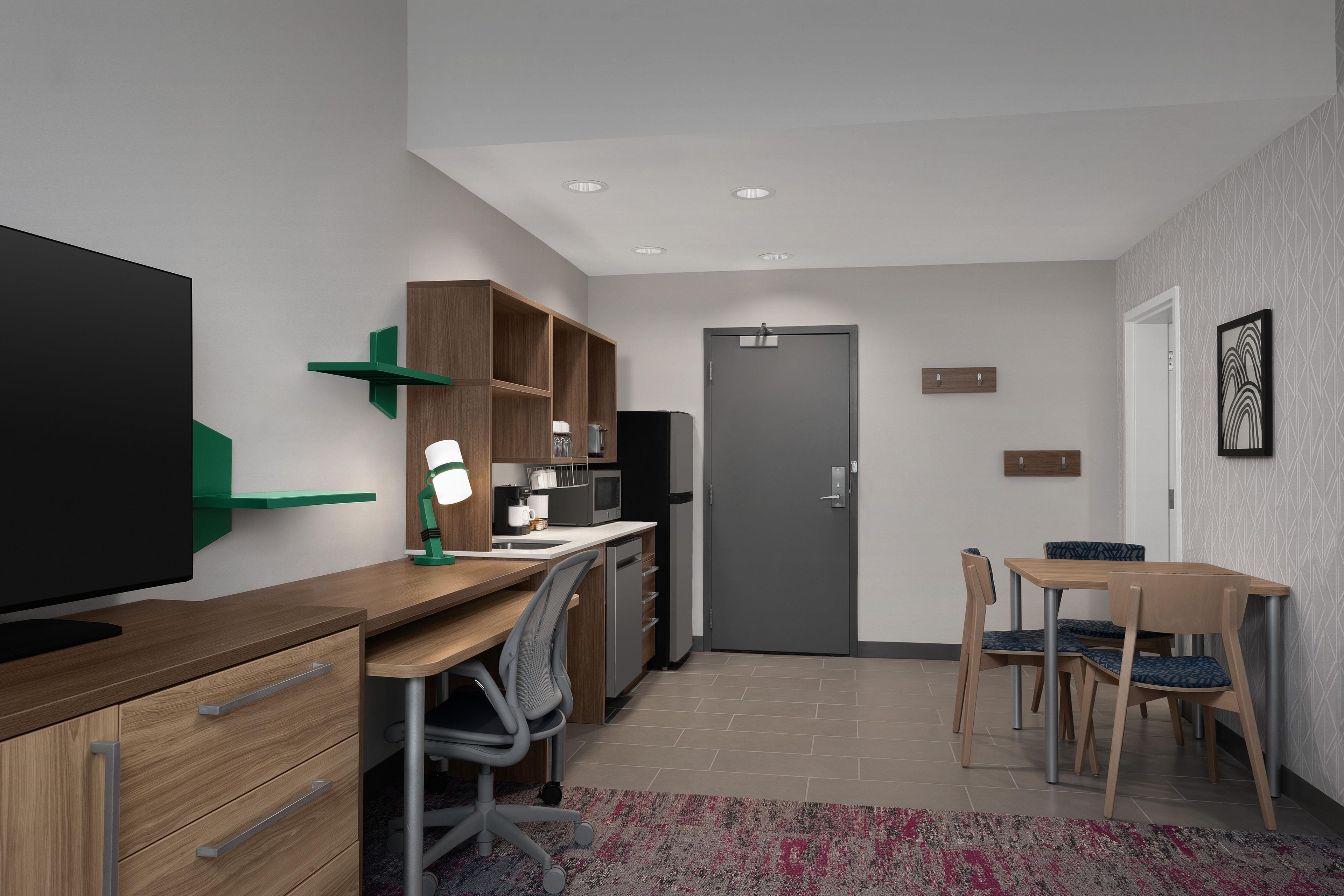 Una habitación de hotel moderna con televisión de pantalla plana, escritorio de madera, silla de oficina, kitchenette y comedor con mesa y sillas.