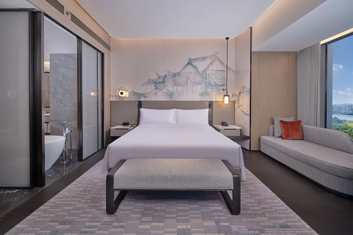 Ein modernes Hotelzimmer mit einem großen Bett, einer Sitzecke, einer Bank, einem Badezimmer mit Waschbecken und Blick auf die Stadt.