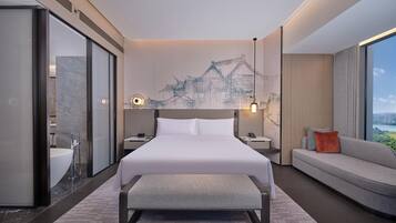 A modern hotel room with a large bed, a sofa, a bench, a bathroom with a sink, and a cityscape view.