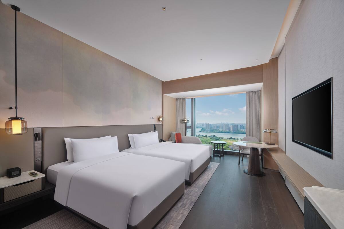 Ein Hotelzimmer mit zwei Betten, einem Fernseher, einem Schreibtisch und Blick auf eine Stadtlandschaft.
