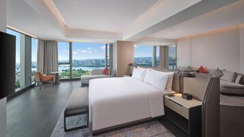 A modern hotel room with a large bed, a desk, and a chair. There is a large window offering a view of a cityscape and a body of water.
