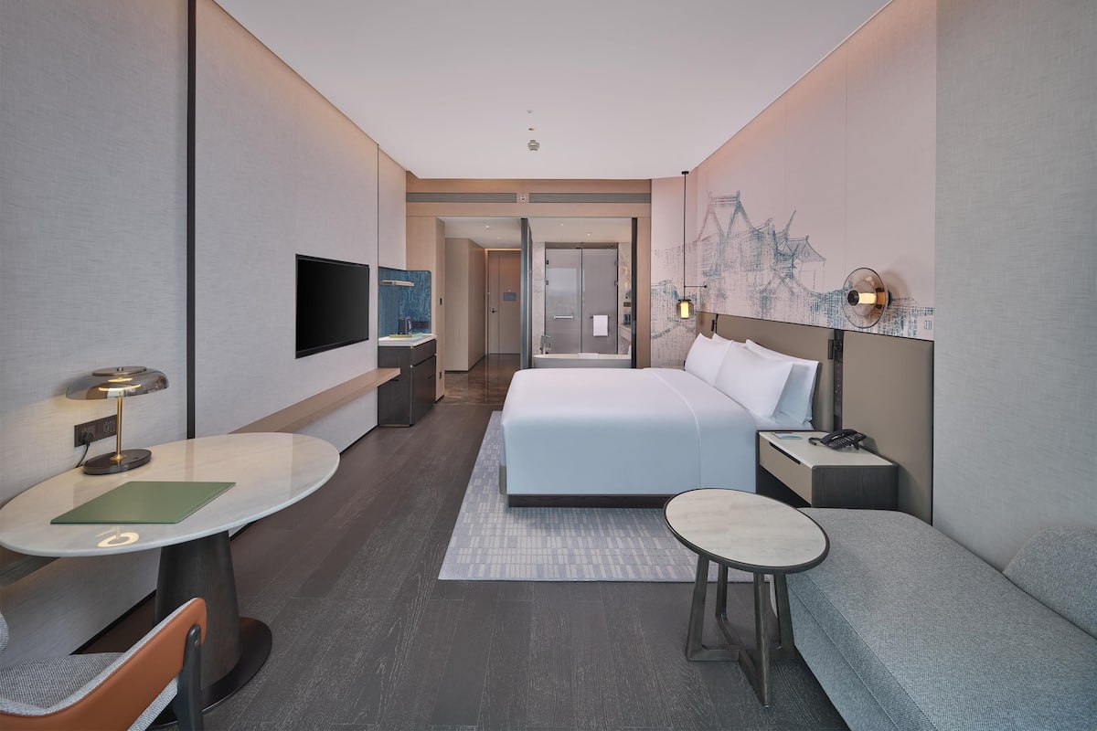 Ein modernes Hotelzimmer mit einem großen Bett, einem Schreibtisch, einem Sessel, einem kleinen Tisch, einem Fernseher und einem Nachttisch.
