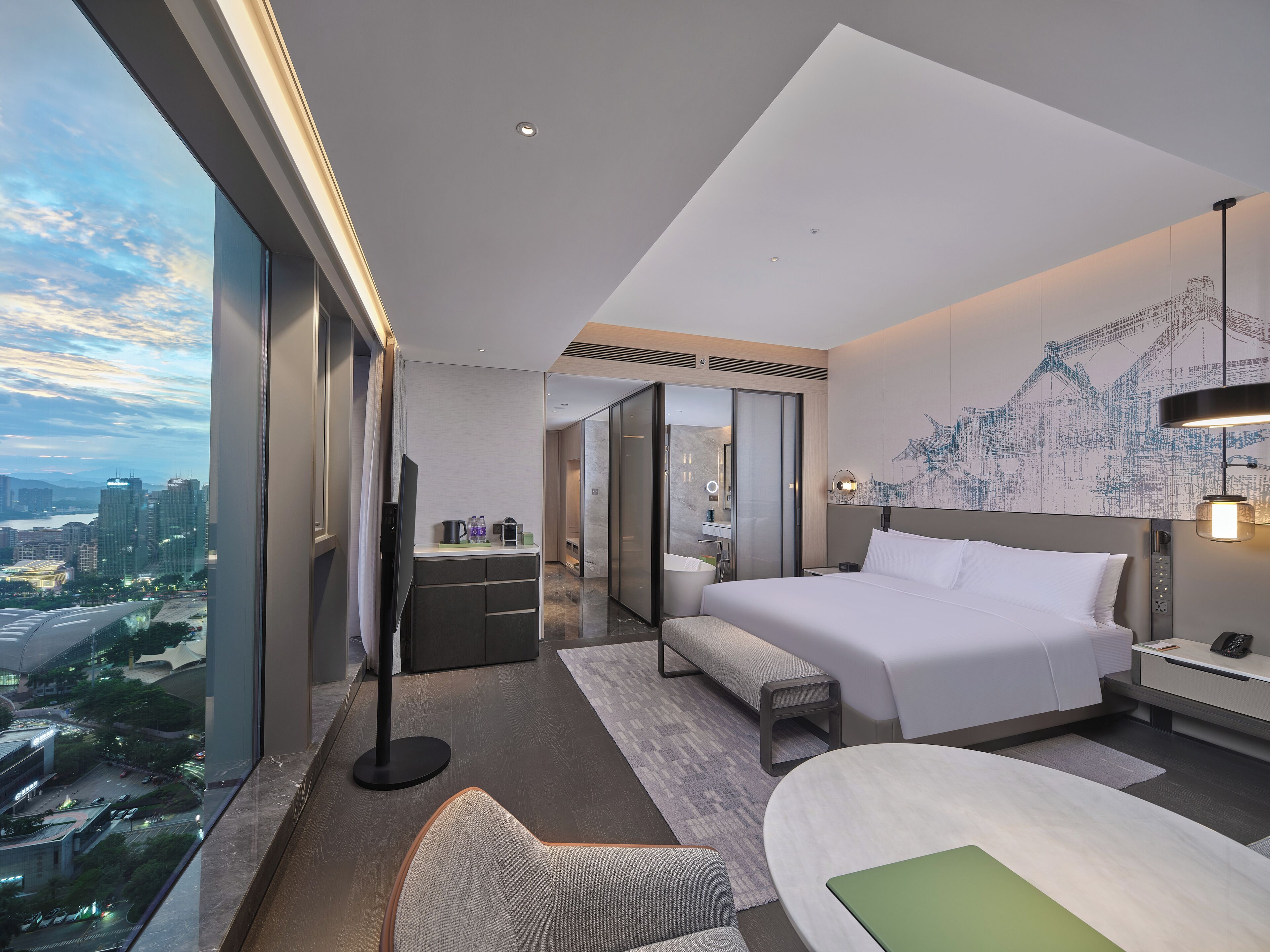 Un cuarto de hotel moderno con una cama grande, un escritorio, una silla, vista a la ciudad y un baño con espejo y lavamanos.