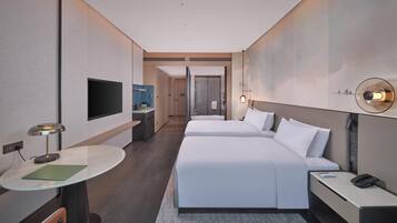 A modern hotel room with two beds, a desk, a chair, a TV, and a bathroom visible in the background.