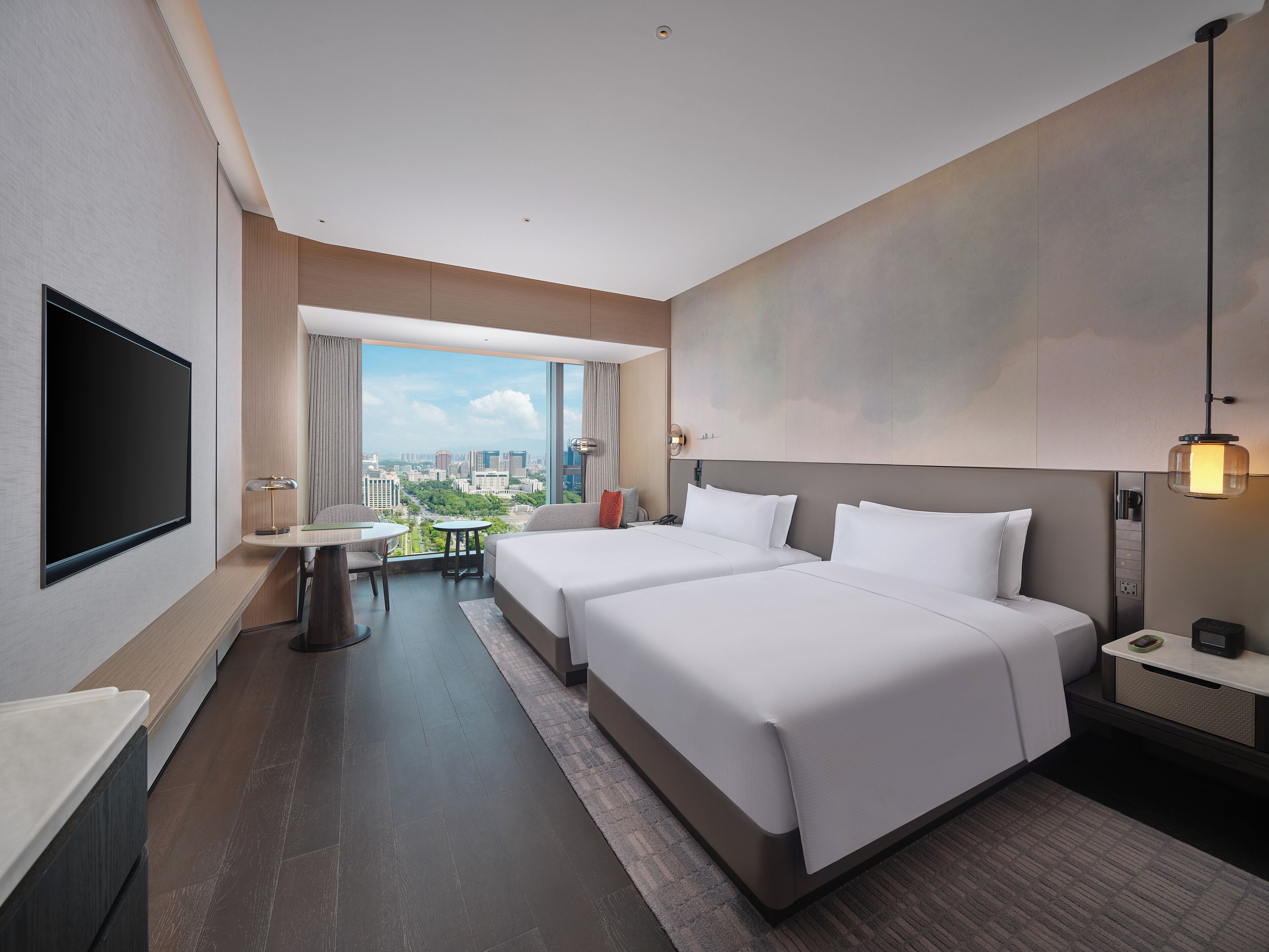 A modern hotel room with two beds, a TV, a desk with a chair, and a large window offering a city view.