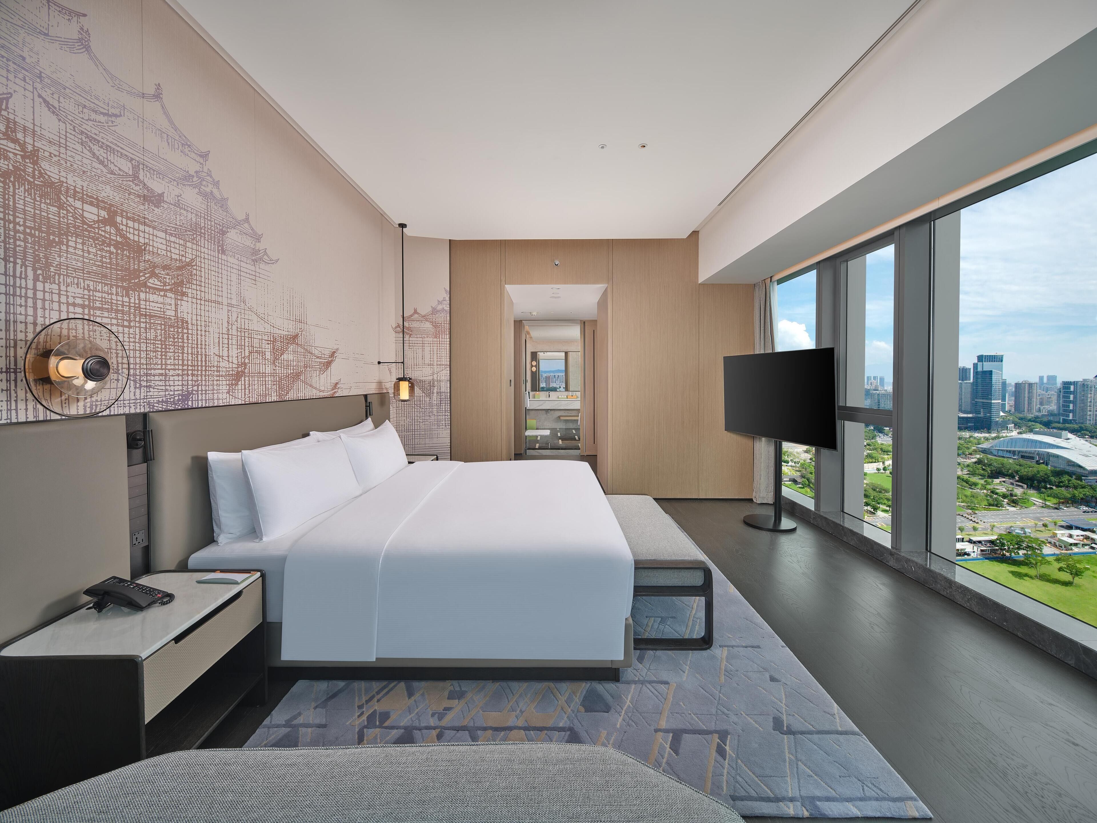 A modern hotel room with a large bed, a desk, and a TV. The room has a city view and a large window.