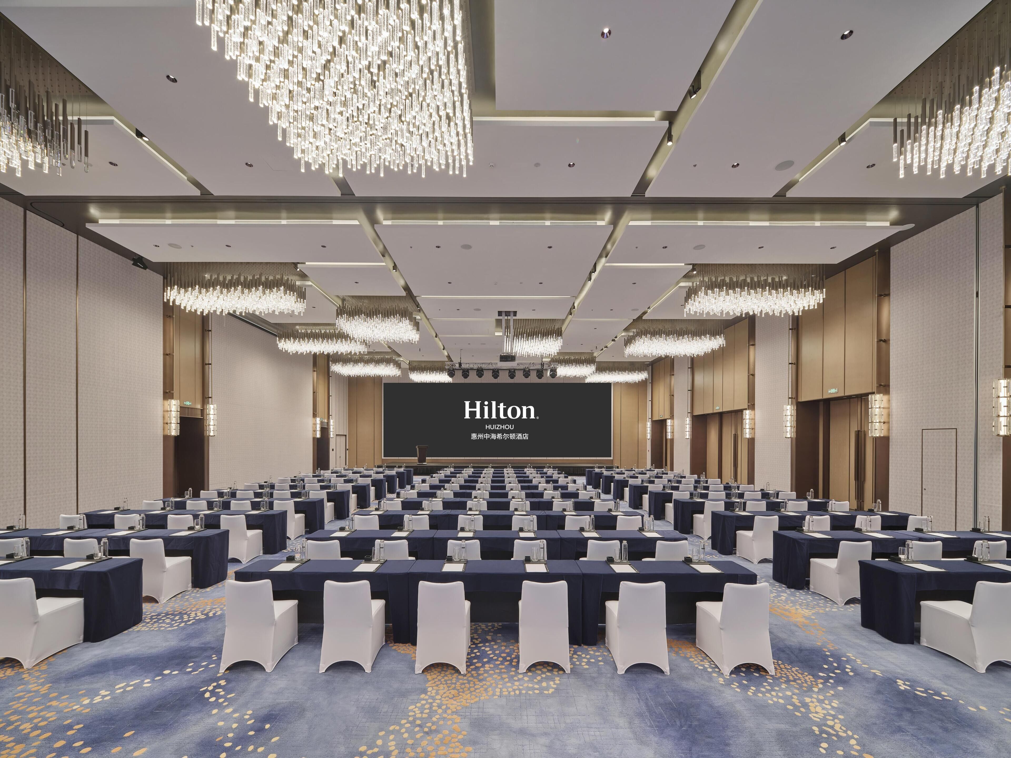 A large conference room with rows of tables and chairs, a large screen displaying 'Hilton', and modern chandeliers.