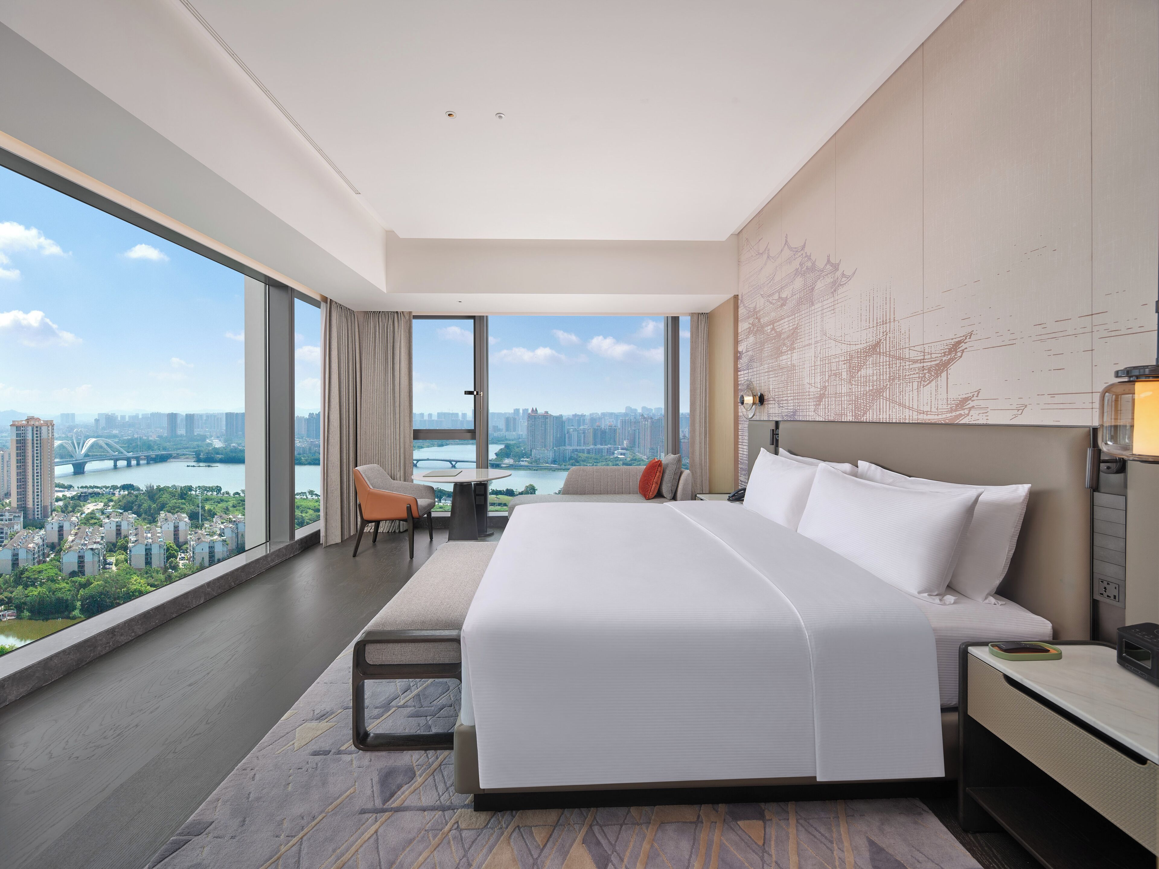 A modern hotel room with a large bed, a desk, and a view of a cityscape through large windows.
