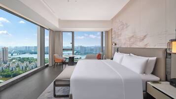 A modern hotel room with a large bed, a desk, and a view of a cityscape through large windows.