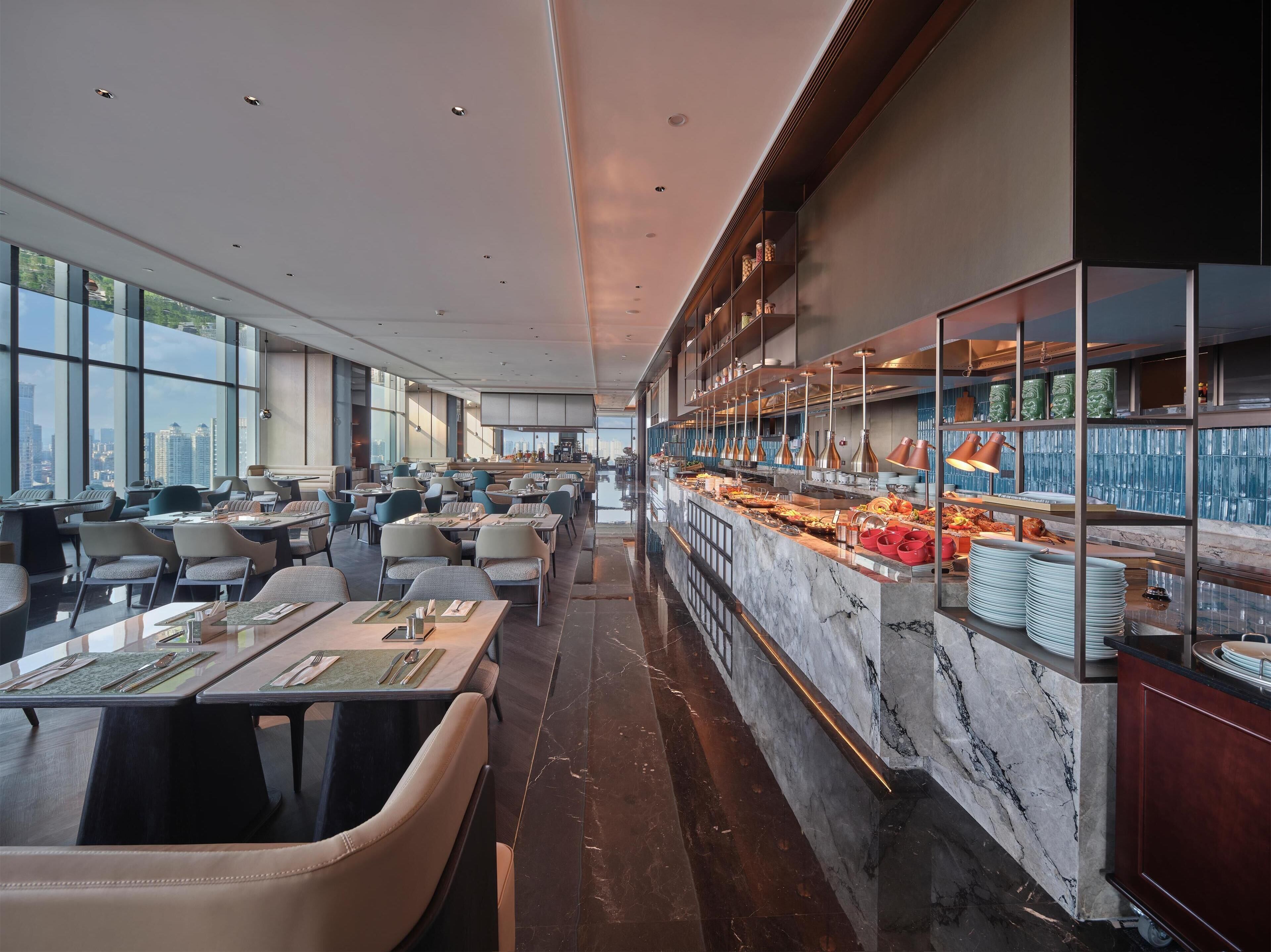 A modern restaurant with a long counter, multiple tables, and a view of the city skyline.