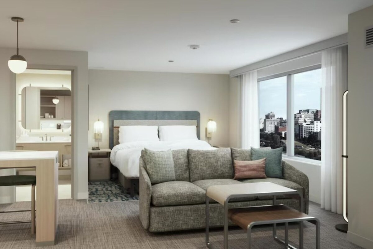 Hotelkamer met een bed, een slaapbank, een eettafel met stoelen, een spiegel en een raam met uitzicht op de stad.