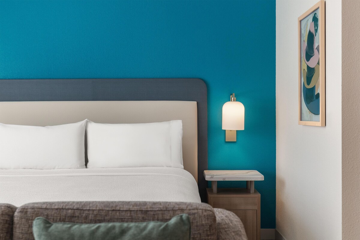 Un lit bien fait, avec du linge de lit blanc, une table de chevet avec une lampe et une œuvre d’art accrochée au mur.