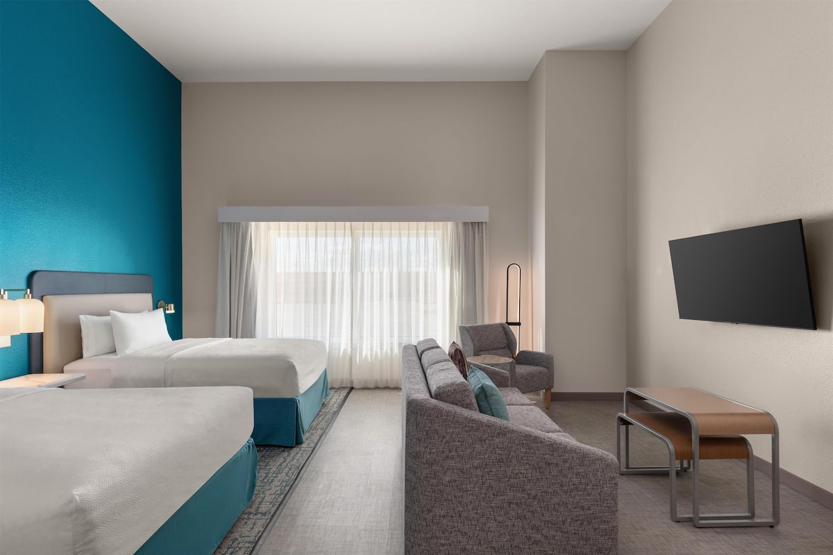 Ein Hotelzimmer mit Bett, Sofa, Ohrensessel, Bank, Fernseher und einem Fenster mit Vorhängen.