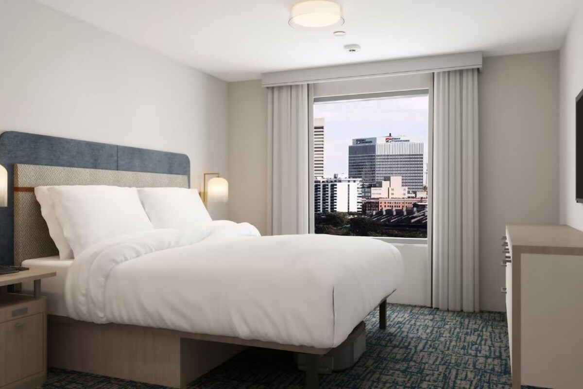 Une chambre d’hôtel équipée d’un lit, d’un bureau, d’une lampe, d’une fenêtre donnant sur la ville et d’un téléviseur.