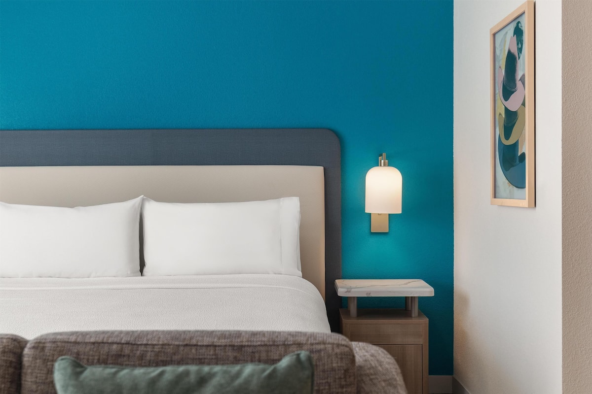 Ein Hotelzimmer mit Bett, Nachttisch, Wandlampe und einem gerahmten Bild.