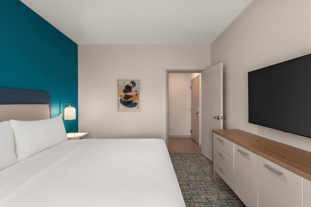Ein Hotelzimmer mit Bett, Nachttisch mit Lampe, an der Wand montiertem Fernseher, einer Tür zu einem weiteren Raum und einem Bild.