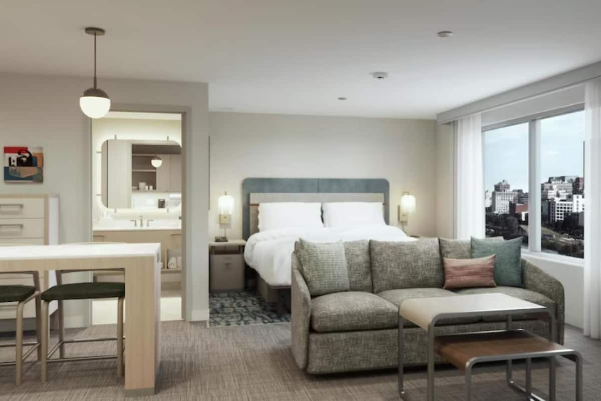 Ein Hotelzimmer mit Bett, einer Couch, einem Esstisch mit Stühlen, einem Spiegel und einem Fenster mit Blick auf die Stadt.