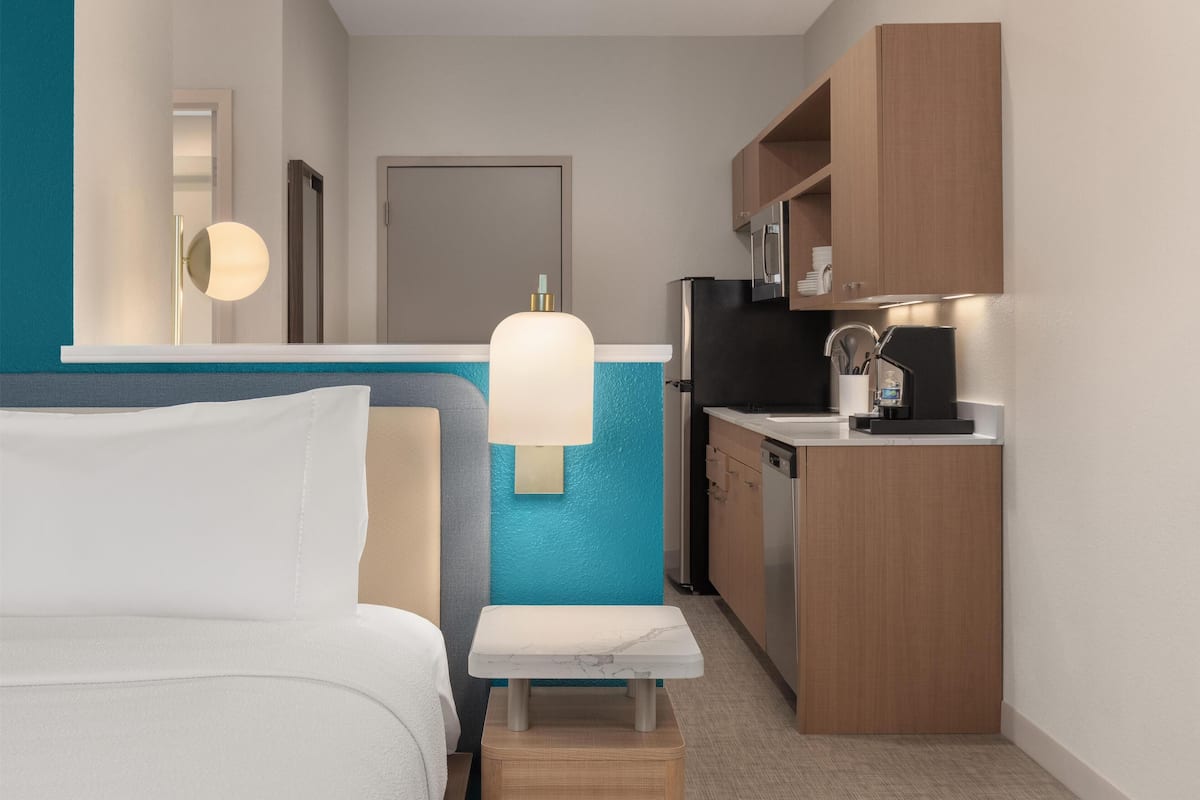 Ein kleines Hotelzimmer mit Bett, Nachttisch, Bank, Küchenzeile und Bad.