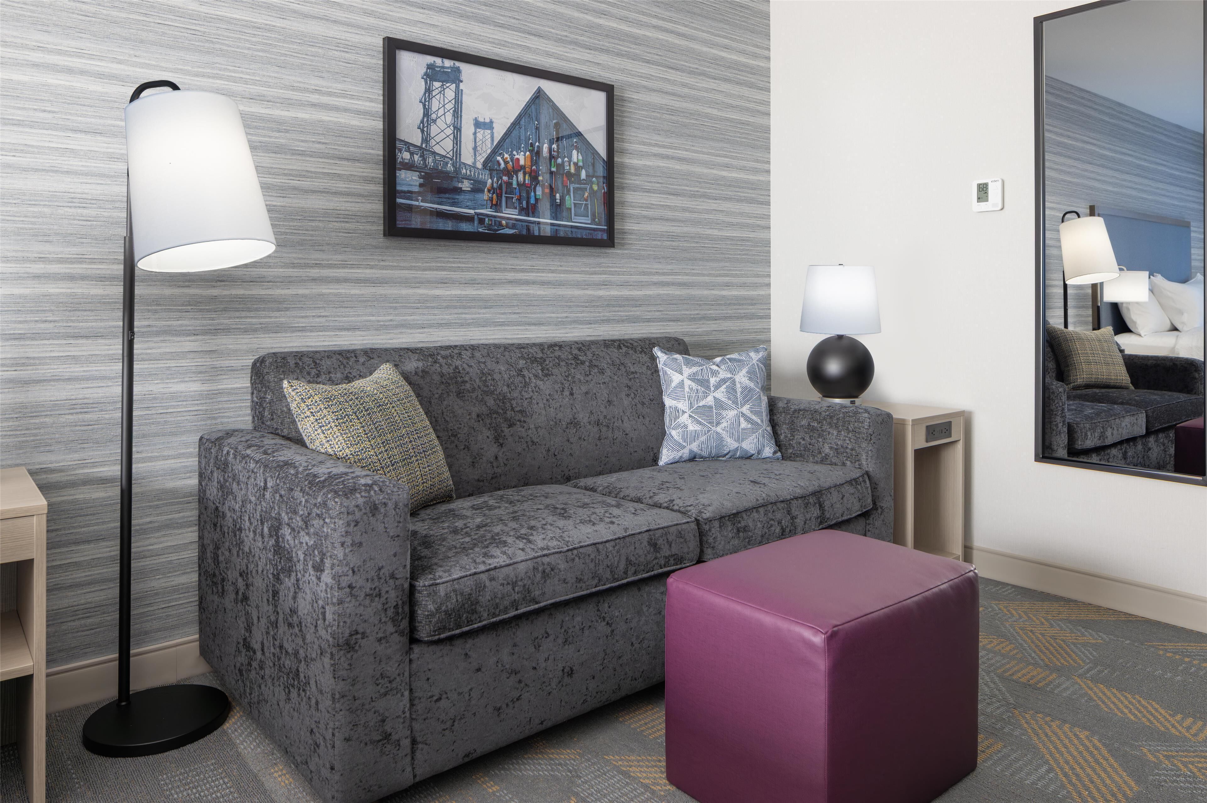 Une chambre d’hôtel moderne avec un canapé gris, un pouf violet, une lampe sur pied, une table d’appoint et une œuvre d’art encadrée au mur.