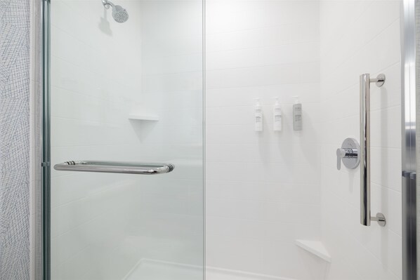 A modern shower area with a glass door, a handrail, and a showerhead. - Home2 Suites By Hilton Kittery (Kittery)