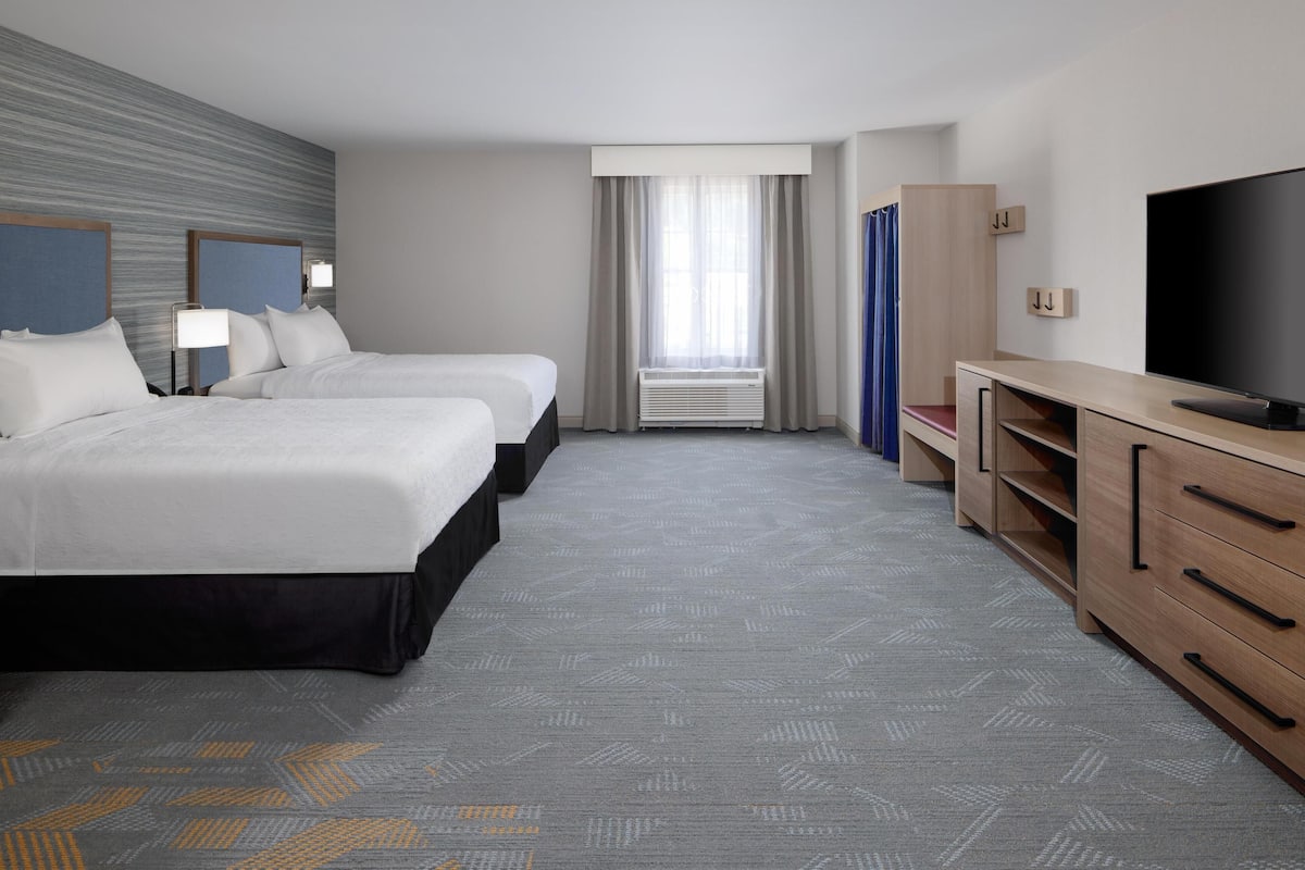 A hotel room with two beds, a TV, a desk, and a large window with curtains.