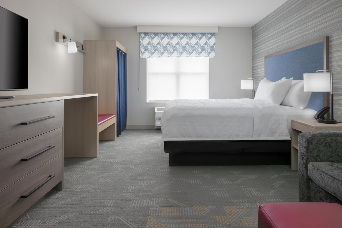 A hotel room with a bed, a TV, a desk, a chair, and a window with a patterned curtain.